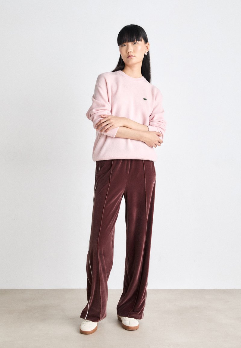 Hellrosa Pullover mit runder Ausschnitt und gesticktem Logo, kombiniert mit burgunderfarbenen Samt-Hosen mit weitem Bein, die weiße Seitenpipings aufweisen.