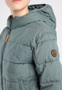 Groene gewatteerde jas met ritssluiting, capuchon en een ronde logopatch op de linkermouw. Soepele textuur met doorgestikt ontwerp.