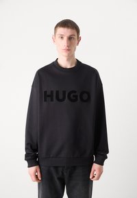 Black sweatshirt featuring a large, raised "HUGO" logo on the front. Made from textured fabric with ribbed cuffs and neckline.