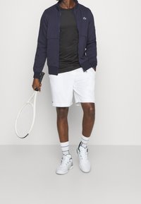 Marinblå zip-up jacka, svart t-shirt, vita shorts, vita träningsskor med accenter. Håller en tennisracket. Randiga vita strumpor.