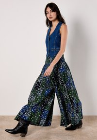Chaleco de mezclilla con botones y un diseño ajustado, combinado con pantalones de pierna ancha con estampado floral en verde, azul y blanco. Botas de tacón negras.