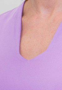 Top a V scollo realizzato in tessuto di colore lilla chiaro, con una texture liscia e un design aderente. Il collo crea una silhouette pulita e semplice.