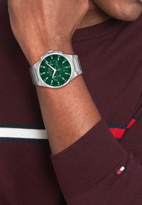 Montre argentée avec un cadran rond vert, des index blancs et des sous-cadrans, sur un bracelet en acier inoxydable. Portée sur un poignet avec un pull bordeaux.