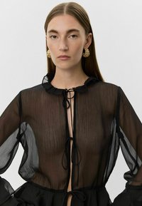 Black sheer blouse with ruffled collar, open front, and tie closure. Features flowing sleeves and textured fabric for a lightweight appearance.