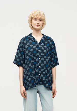 Person med krøllete blondt hår har på seg en marineblå kortermet skjorte med hvit og blå geometrisk mønster og lyseblå jeans.