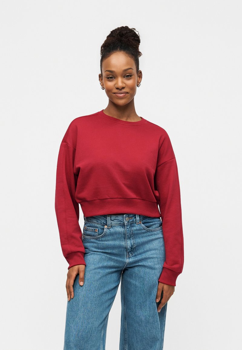 Young woman with hair in a bun wearing a red long-sleeve cropped sweatshirt and high-waisted blue jeans, standing against a plain light background.