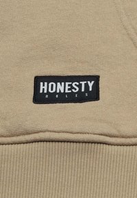 Tan Hoodie Material mit gerippter Textur, versehen mit einem schwarzen rechteckigen Aufnäher, auf dem in fetten weißen Buchstaben "HONESTY RULES" steht.