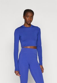 Mėlynas ilgomis rankovėmis trumpas viršus su apvalia iškirpte ir prigludusiomis rankovėmis, derinamas su aukštu liemeniu mėlynomis legging'omis, turinčiomis struktūrizuotus siūlus.