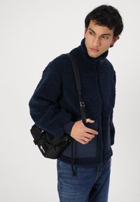 Marineblaue Fleecejacke mit hohem Kragen, Reißverschluss und strukturiertem Aussehen. Kombiniert mit einer schwarzen Umhängetasche und blauen Jeans.