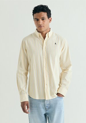 Mann trägt hellgelbes und weiß gestreiftes Hemd mit Knöpfen und hellblaue Jeans vor einfarbigem Hintergrund.