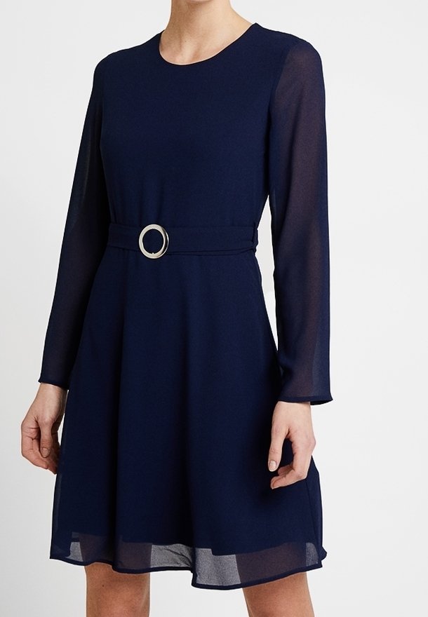 Femme portant une robe bleu marine à manches longues avec un décolleté rond et une ceinture avec une boucle métallique circulaire à la taille.