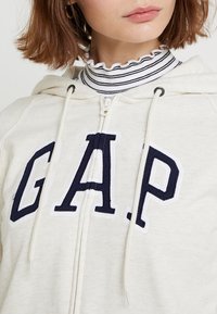 Sudadera con cremallera de color crema con el logotipo bordado en azul marino "GAP", que cuenta con cordones y una camisa tipo polo blanca y azul marino de rayas texturizadas debajo.