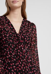 Blouse noire à motifs floraux avec manches longues et transparentes. Présente un décolleté en V profond et des motifs floraux rose et rouge complexes sur le tissu.