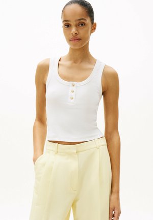 Top court blanc à encolure large avec trois accents de boutons dorés à l'avant. Texture lisse et côtelée. Associé à un pantalon jaune clair.