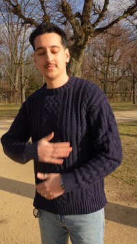 Jeune homme en pull bleu marine et jean debout dans un parc avec des arbres dépouillés, les mains posées sur son ventre et les yeux fermés.