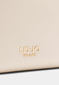 Logo Liu Jo Milano na textúrovanej, svetlej béžovej koženej ploche so zlatými písmenami, s hladkým povrchom a jemným vzorom zrna.
