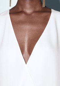 Abito avvolgente bianco con scollatura a V profonda, tessuto liscio e orecchini statement dorati. La scollatura mette in risalto la zona della clavicola.