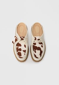 Un paio di scarpe beige senza lacci con motivo di pelle di mucca marrone e bianca sulla parte superiore e suole in gomma testurizzata color miele, viste dall'alto.