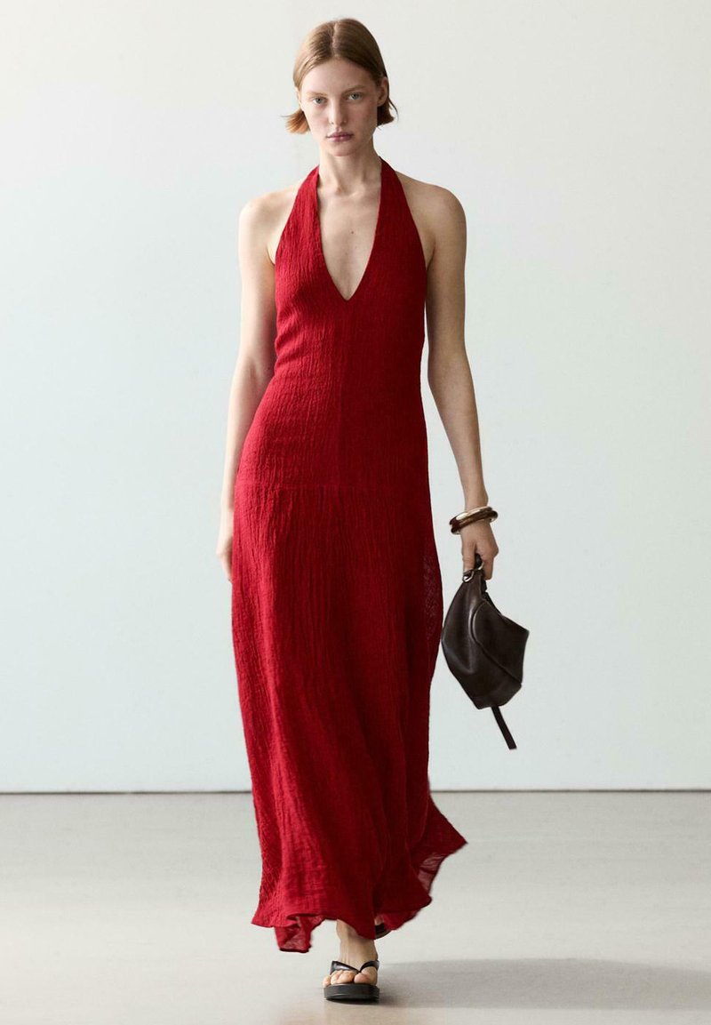 Vrouw die loopt, gekleed in een mouwloze rode halter maxi-jurk, een kleine zwarte handtas vasthoudend, met een neutrale achtergrond.