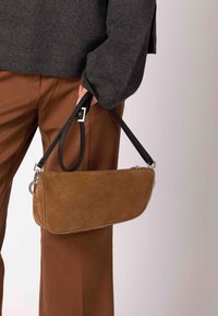 Brun ruskind clutch taske med en sort læderrem, der har en sølvfarvet metaldekoration og et præget brandlogo på forsiden.