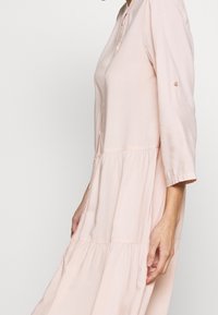 Abito rosa chiaro a maniche lunghe con scollo a bottoni, design a strati e lievi arricciature in vita. Tessuto morbido.