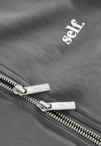 Gray fabric zip-up jacket with silver zippers, featuring "self." branding on the left side and smooth texture. Zippers have tags embossed with "self."