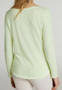 Žena v světle žluté dlouhé košili a světle béžových kalhotách, zobrazená zezadu na jednobarevném pozadí.