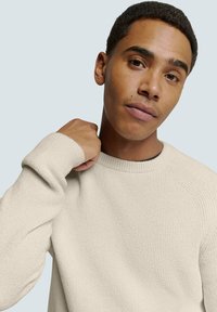 Crème-kleurige geribbelde trui met een ronde hals, textuurstof en ingesette mouwen. Model poseert met hand op de nek tegen een lichte achtergrond.