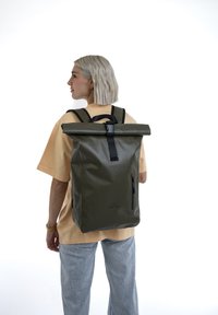 Olíva zöld, vízálló anyagból készült, felgöngyölhető hátizsák, állítható fekete pántokkal és oldalsó cipzáras zsebbel.