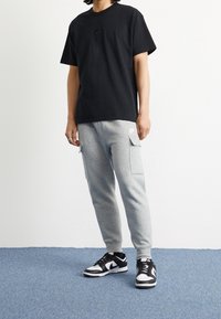 Svart bomulls t-shirt med en liten svart logotyp, kombinerad med grå cargobyxor i sweatpantsmodell med sidofickor, och svart-vita sportskor.