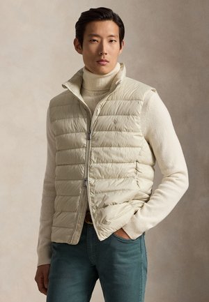 Mężczyzna w kremowej pikowanej kamizelce na kremowym swetrze z golfem i niebieskich dżinsach, stojący na jednolitym neutralnym tle.