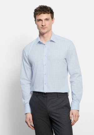 Mann trägt hellblau kariertes Hemd mit Knöpfen und dunkelgraue Hose, steht vor schlichtem weißem Hintergrund.