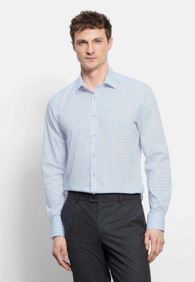 Homme portant une chemise à carreaux bleu clair boutonnée et un pantalon gris foncé, debout devant un fond blanc uni.