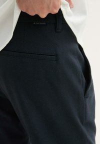 Navy textured pants with a slim fit, featuring a discreet logo label, side and back pockets. Made of woven fabric with a subtle pattern.