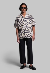 Camicia a maniche corte con bottoni di colore beige con strisce di tigre nere, abbinata a pantaloni neri a gamba larga e scarpe nere, caratterizzate da un motivo testurizzato.