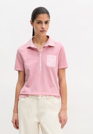 Kobieta o ciemnych włosach związanych w kucyk, ubrana w różową koszulkę polo z krótkim rękawem i kremowe spodnie, na gładkim białym tle.