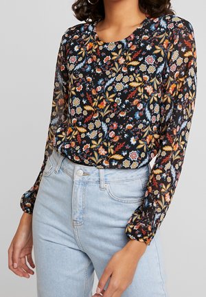 Vrouw draagt een zwarte blouse met een kleurrijke bloemenprint en doorschijnende lange mouwen, gecombineerd met lichtblauwe high-waisted jeans.