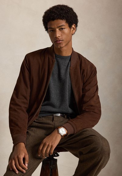 Giovane uomo con capelli ricci che indossa una giacca marrone, un maglione grigio, pantaloni a quadri e un orologio, seduto davanti a uno sfondo beige semplice.