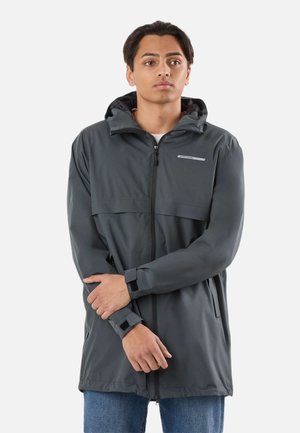 Ung mand iført en mørkegrå hættejakke, der er vandtæt, med justerbare remme ved håndleddene og lynlås foran, stående med armene krydset.