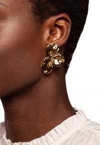Orecchini floreali in tonalità oro con una finitura texturizzata, che assomigliano a petali, attaccati a un piccolo cerchio. Indossati su una tonalità di pelle texturizzata.