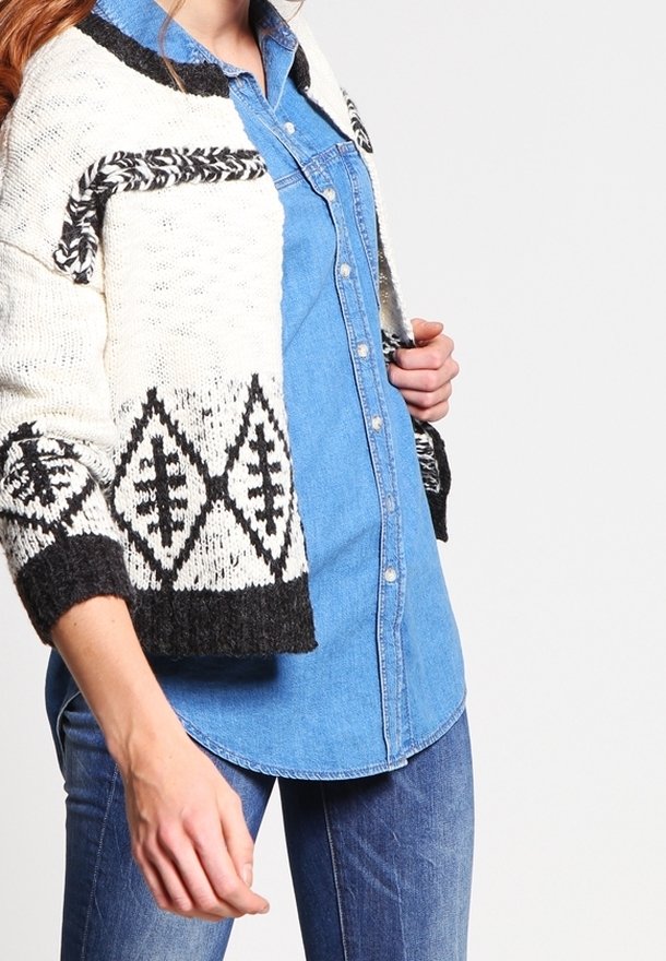 Femme portant un cardigan en tricot à motifs noirs et blancs par-dessus une chemise en denim bleu boutonnée et un jean bleu, vue du cou jusqu'aux hanches.