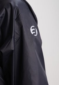 Veste imperméable noire avec un emblème circulaire blanc contenant les lettres stylisées "E" et "J" dans le dos.