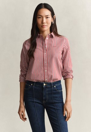 Femme aux longs cheveux foncés portant une chemise boutonnée rayée rouge et blanche et un jean bleu foncé, debout devant un fond uni.