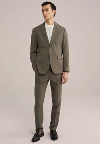 Olive zöld öltöny, illeszkedő blézerrel és sima frontrésszel rendelkező nadrággal, texturált anyaggal, kivágott hajtókákkal és barna gombokkal. Fehér inggel párosítva.