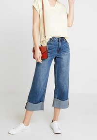 Lysegul kortærmet top, blå brede cropped jeans med splittersøm, hvide sneakers, rød crossover-taske, minimale accessories.