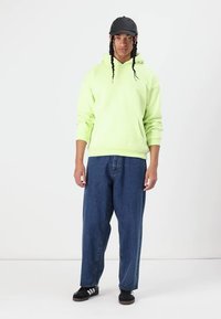 Grøn bomulds-hættetrøje med kængurulomme, parret med løse blå jeans og sorte sneakers. Tilbehøret inkluderer en sort kasket.