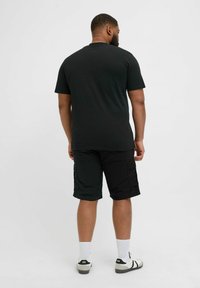 Schwarzes Kurzarm-T-Shirt aus Baumwolle, kombiniert mit schwarzen Cargo-Shorts mit Seitentaschen, getragen mit weißen Socken und schwarzen Schuhen.