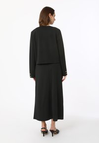 Ensemble noir composé d'une veste courte à manches longues et d'une jupe longue. La veste est ornée de boutons dorés aux poignets.