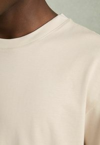 Tricou din bumbac de culoare crem, cu gât rotund, mâneci scurte și o textură ușoară, netedă. Detalii minime de coasere vizibile.