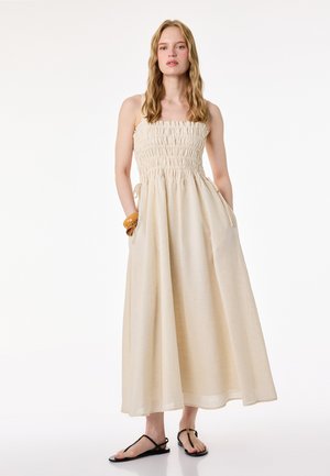 Femme debout portant une robe maxi beige sans manches avec un corsage froncé, des sandales plates noires et un bracelet en bois au poignet.